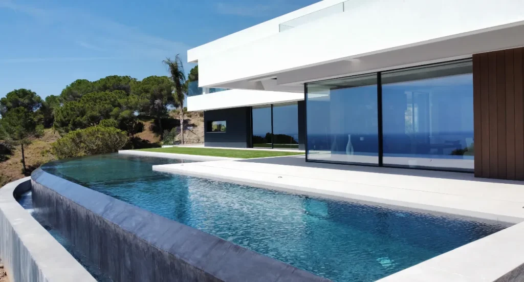 Piscine à débordement incurvé devant une villa contemporaine, avec vue sur les pins et le ciel bleu.