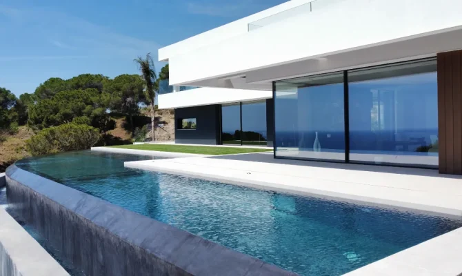 Piscine à débordement incurvé devant une villa contemporaine, avec vue sur les pins et le ciel bleu.